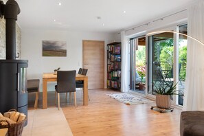 A modern interior with a wood-burning stove, a dining area with chairs, a bookshelf, and a view through a glass door to an outdoor area with plants and a spiral staircase.