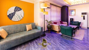 A modern hotel lobby with a grey sofa, a round wall art piece, a floor lamp, and a seating area with a patterned rug. - IKE CITY Hotel (Tema)