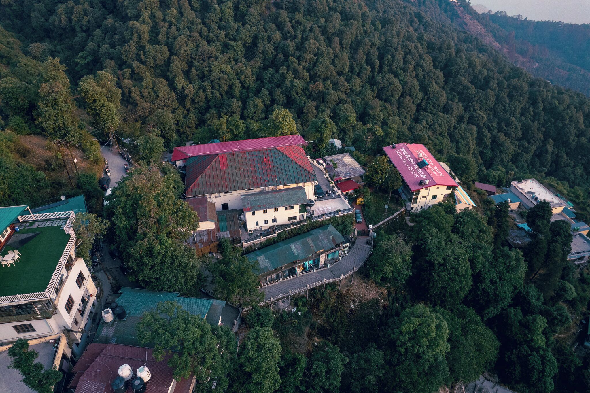 An aerial view of a building complex surrounded by dense forest, featuring multiple structures with red roofs and a green lawn area.
