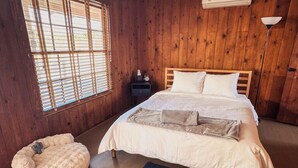 Ein Zimmer mit Holzboden, einem Bett, einem Nachttisch, einer Lampe und einem Fenster mit Jalousien.