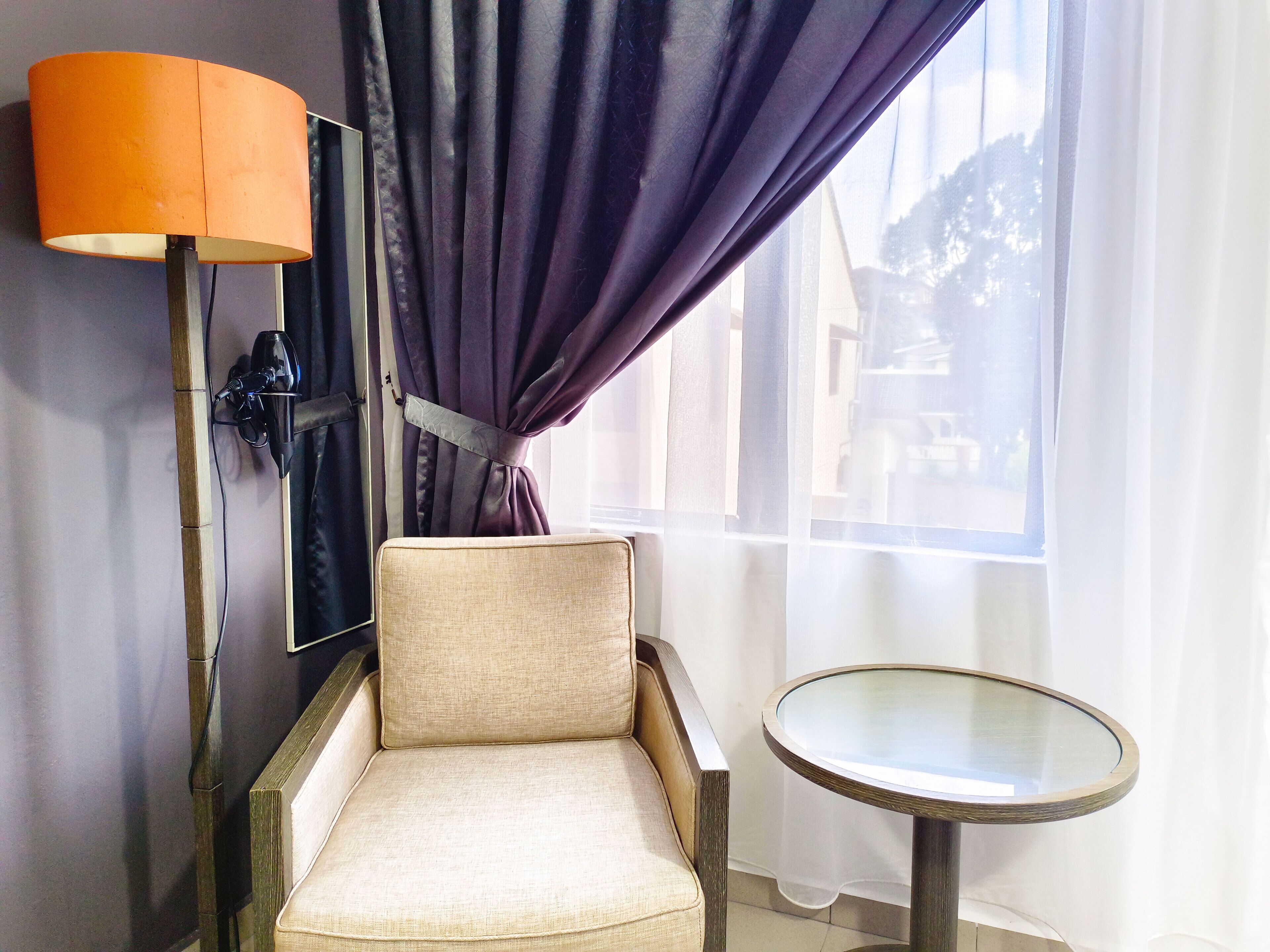 A room with a beige armchair, a small round table, a lamp, and a window with purple curtains.