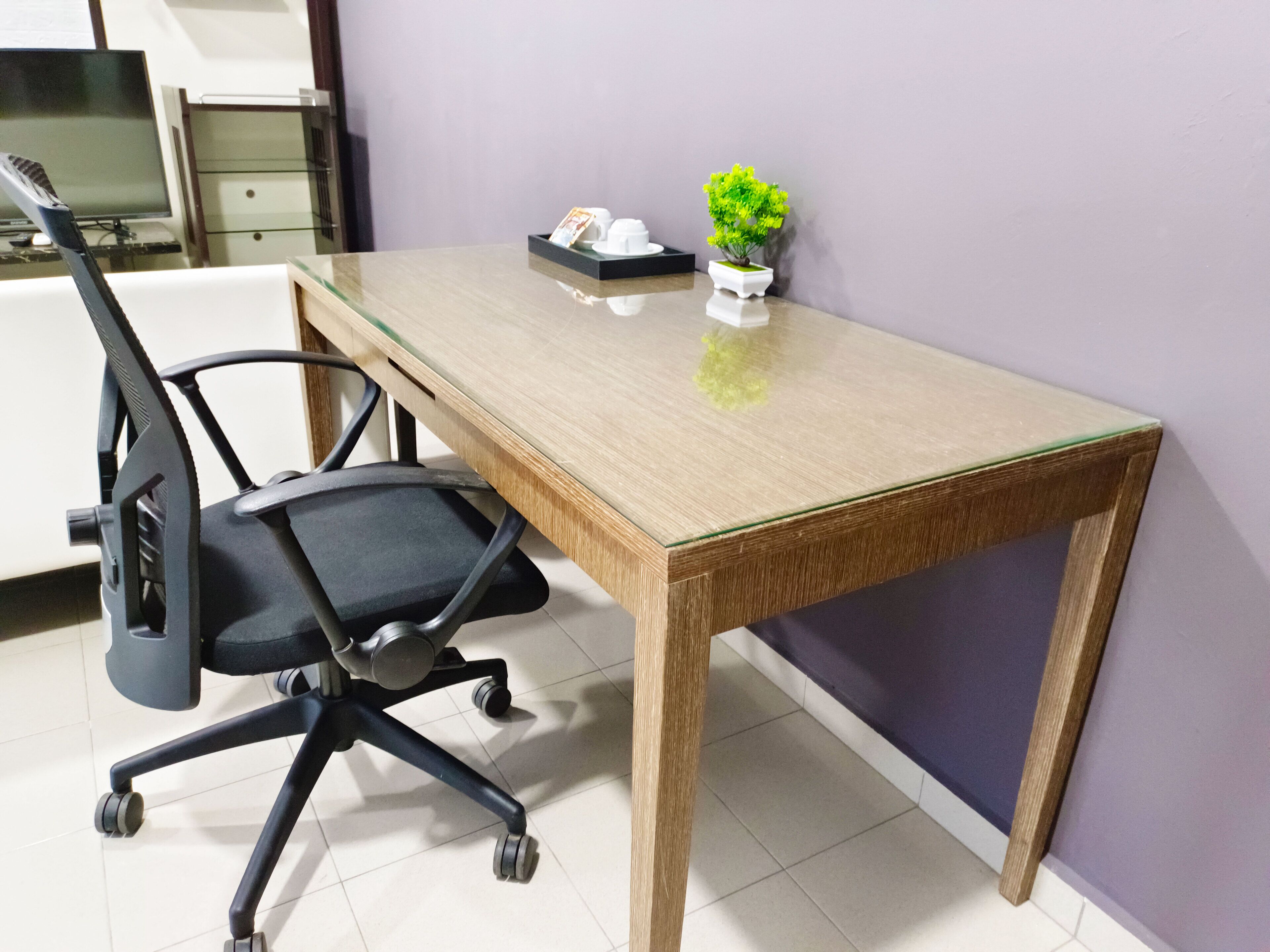 A wooden desk with a glass top, a black office chair, a small potted plant, and a white object on the desk.