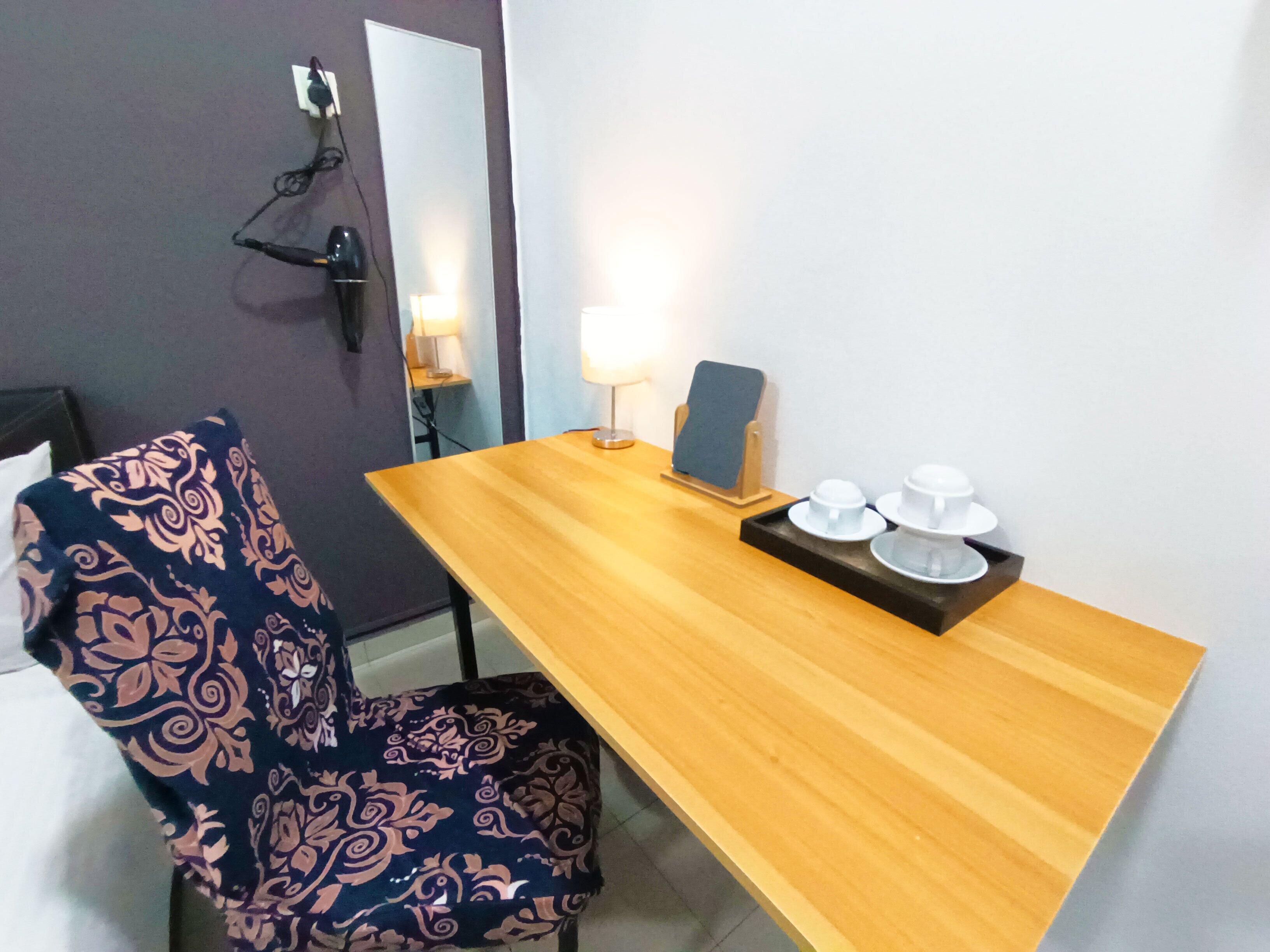 A room with a wooden desk, a chair, a mirror, and a bedside table with a lamp and cups.