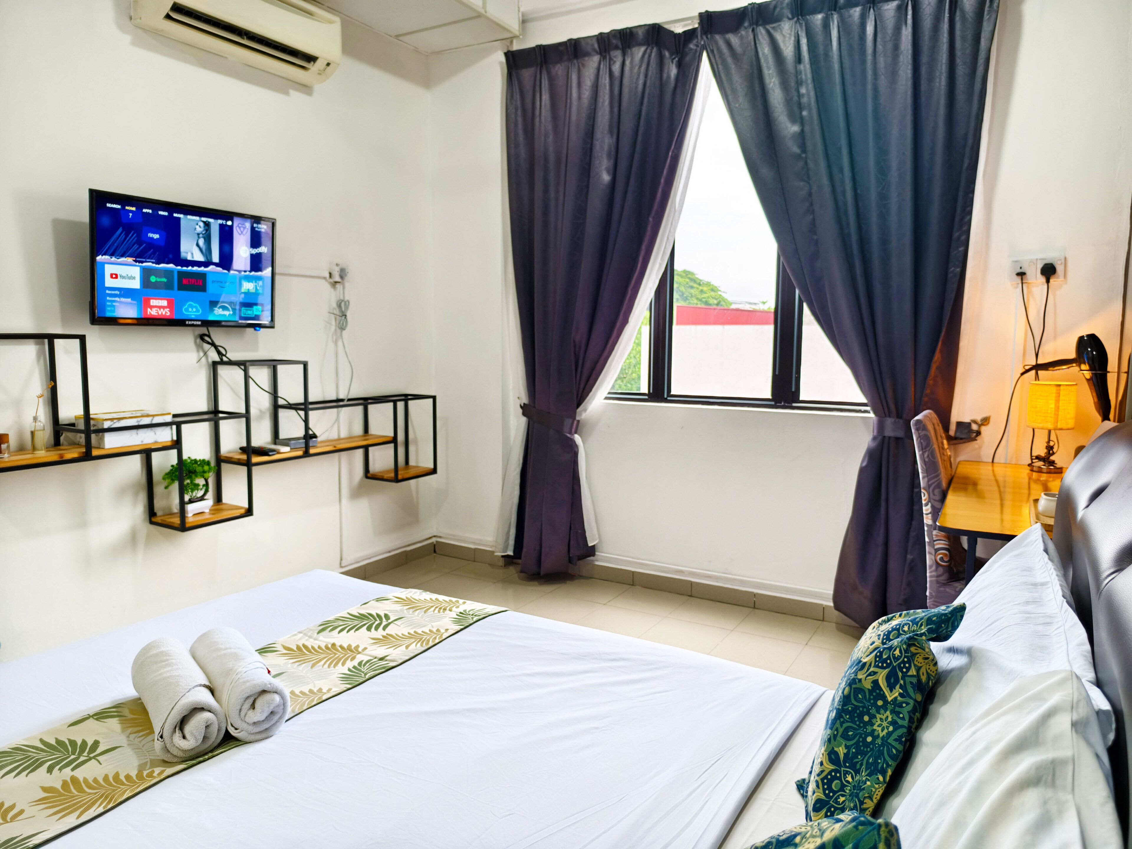 A hotel room with a bed, a TV mounted on the wall, a desk with a lamp, and a window with curtains.