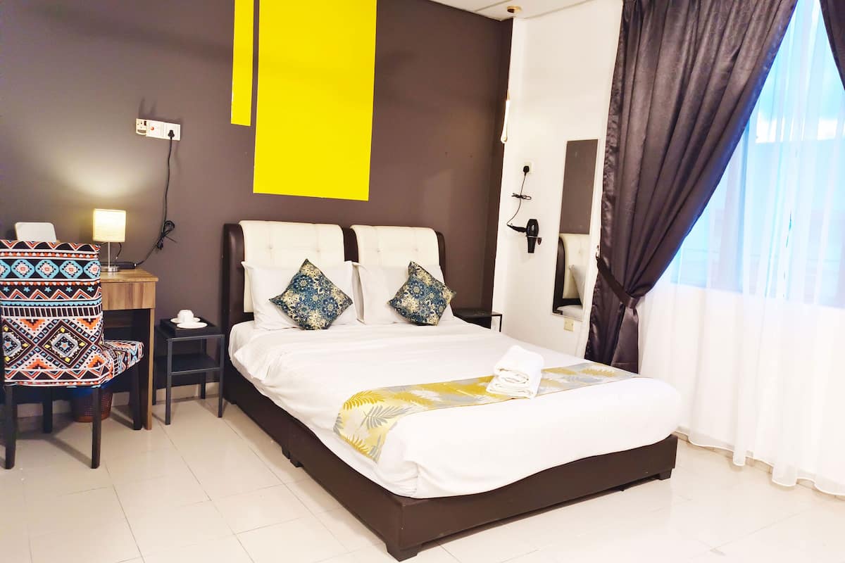 Une chambre d’hôtel avec un lit, un bureau, une chaise, une lampe et une grande fenêtre avec des rideaux.
