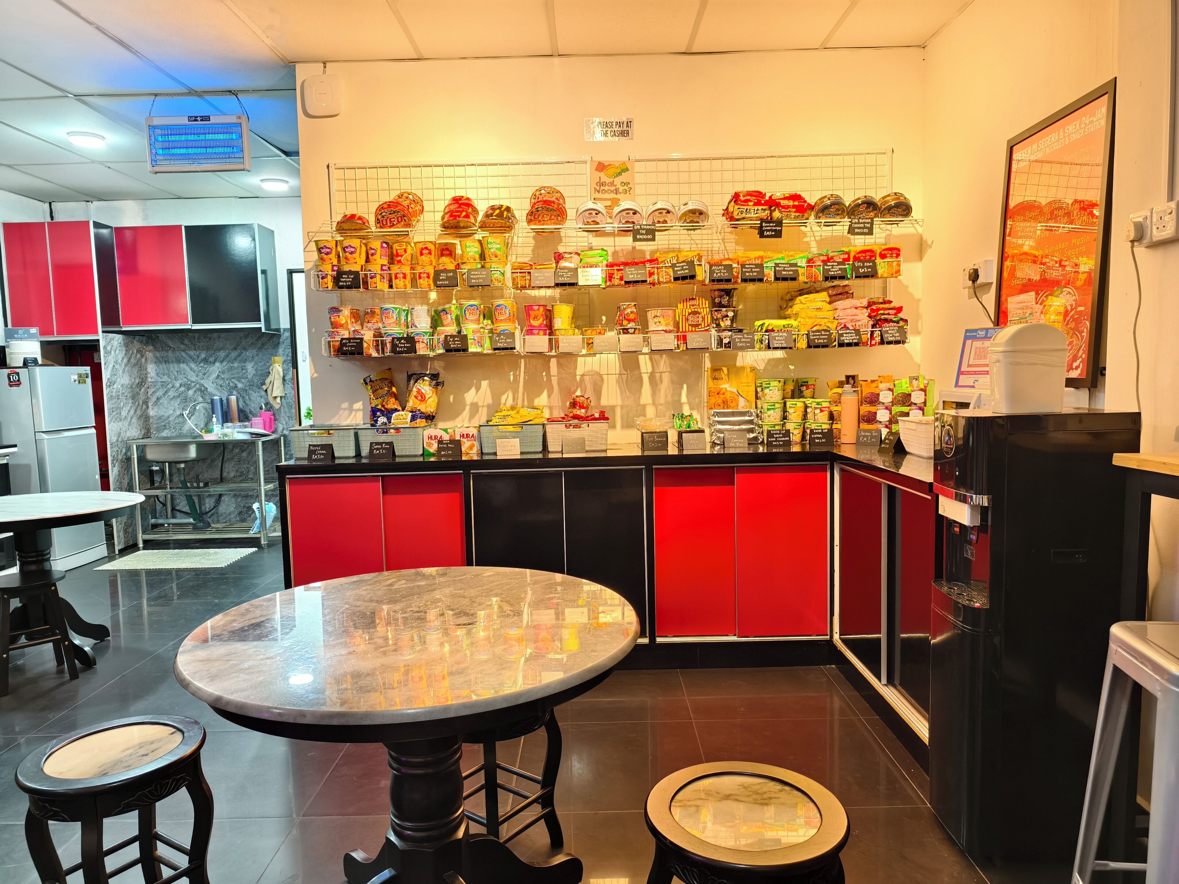 Interior de un restaurante con un mostrador que exhibe diferentes alimentos, una máquina de café y una zona de estar con mesas y taburetes.