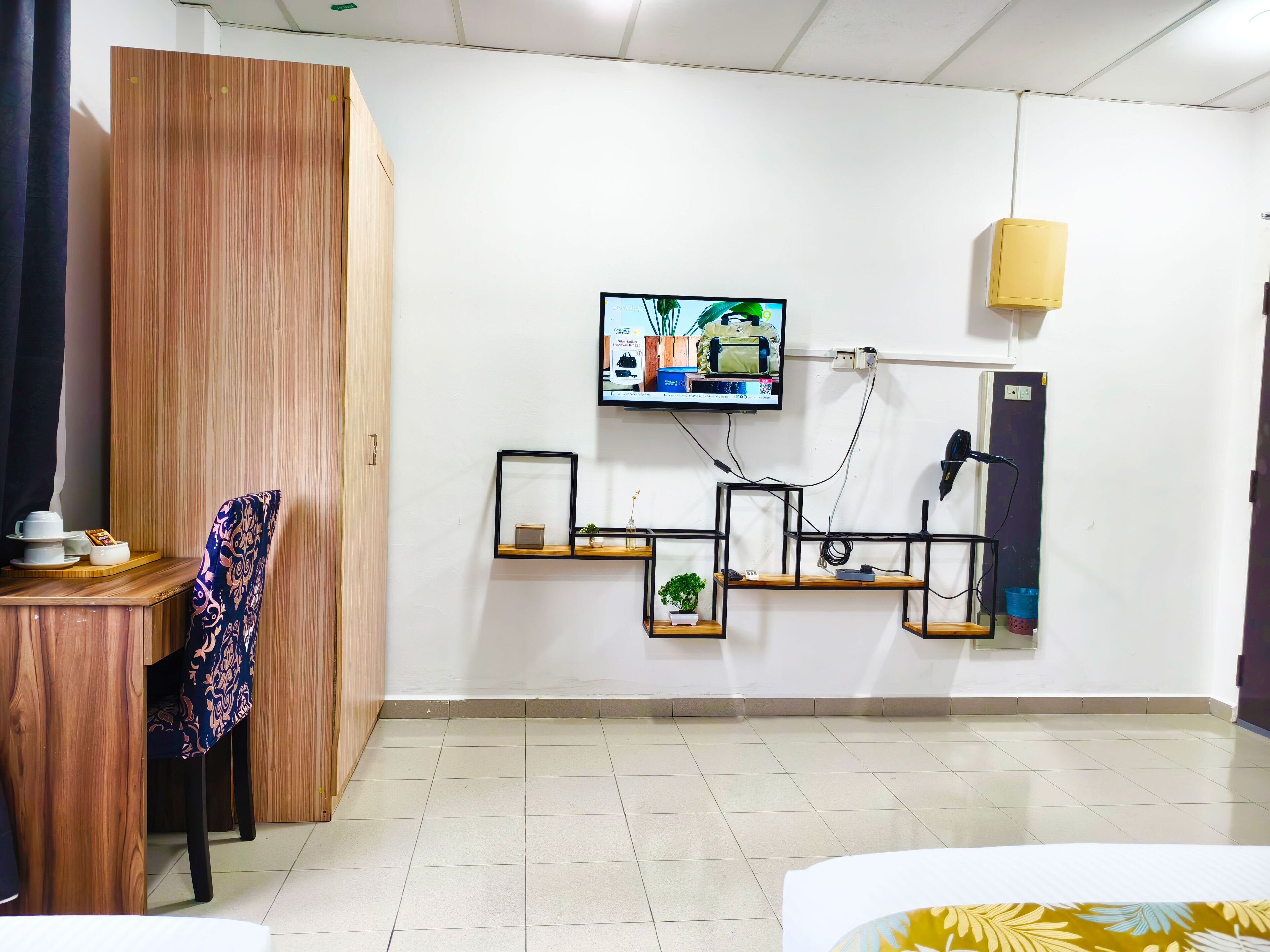 A modern hotel room with a wooden wardrobe, a desk with a chair, a mounted TV, and a wall-mounted shelf with a plant.