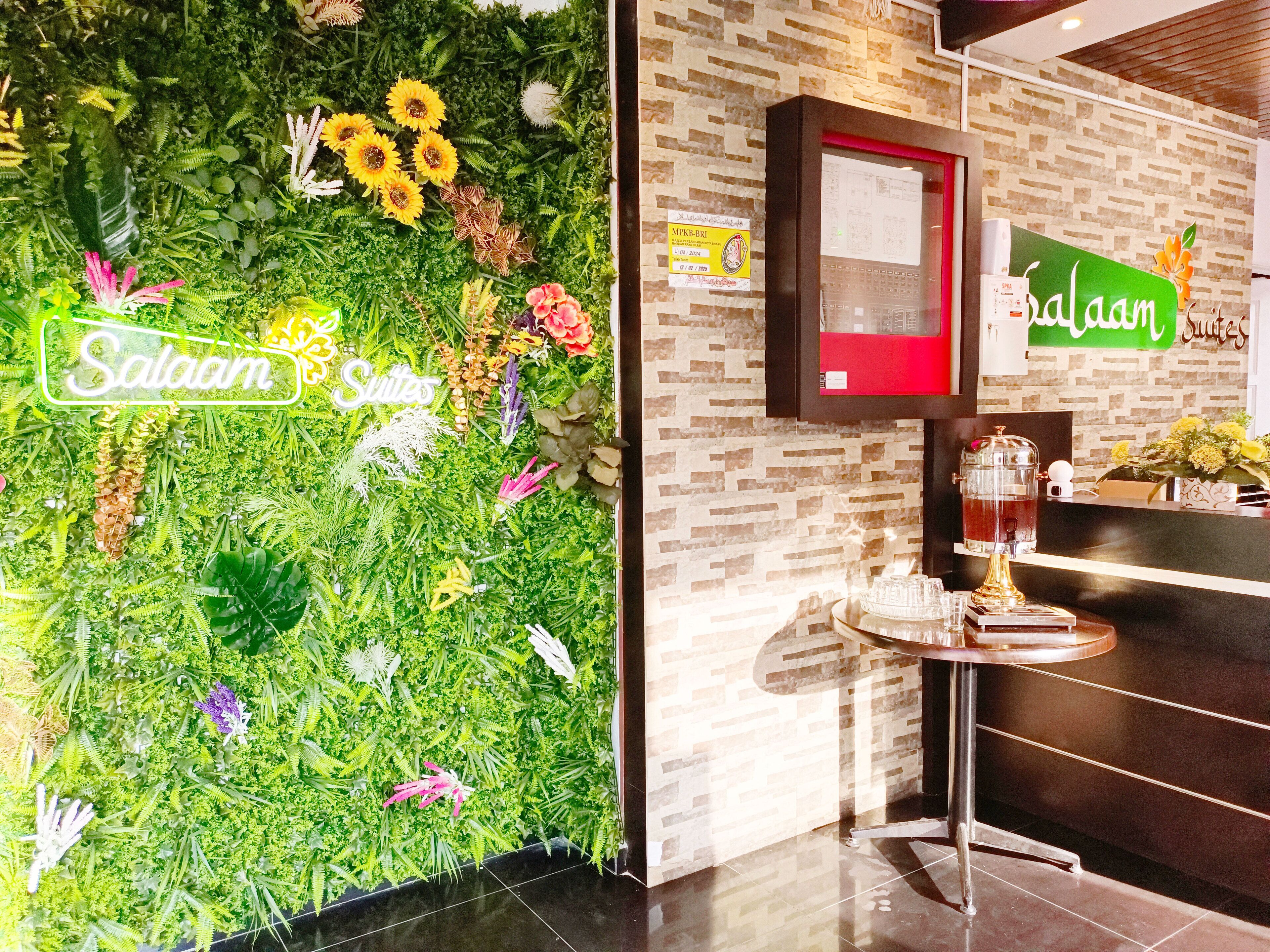 Un área de recepción con una pared verde decorada con plantas y flores, un mostrador de recepción y un cartel que dice "Salaam Suites".