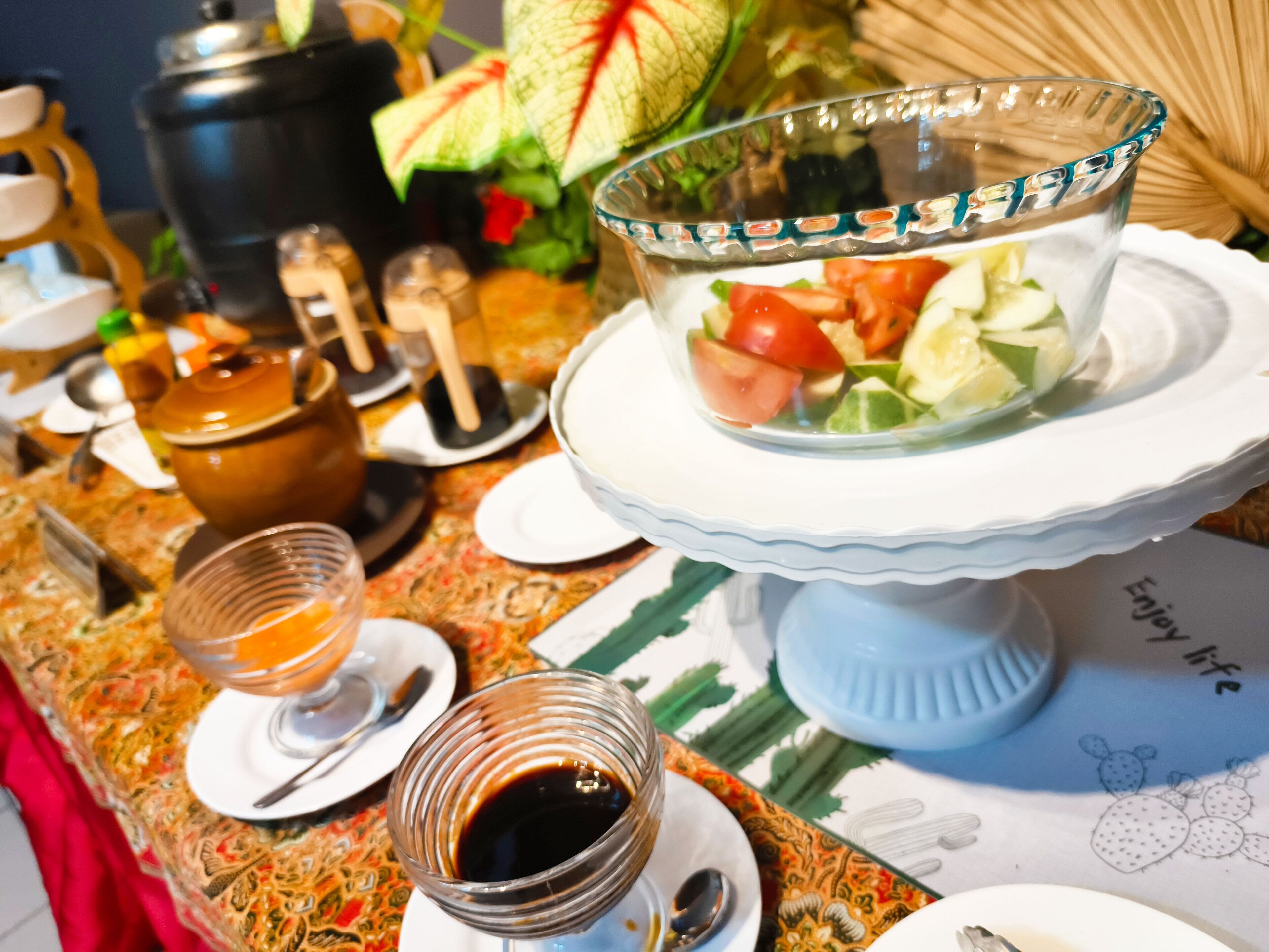 A breakfast setting with a fruit salad, tea, and a coffee pot on a patterned tablecloth.