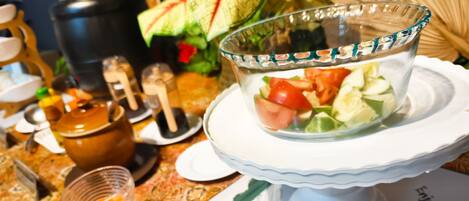 A breakfast setting with a fruit salad, tea, and a coffee pot on a patterned tablecloth.