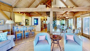 A spacious living area with wooden beams, a dining area with wicker chairs, and a kitchen visible in the background. - Seaglass - Waterfront, Incredible Views of BHI Creek, 3 Suites, Pool, Kayaks, BHI Club+Golf, Dogs OK (Bald Head Island)