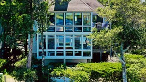 A multi-story house with large windows and a balcony, surrounded by lush greenery. - Seaglass - Waterfront, Incredible Views of BHI Creek, 3 Suites, Pool, Kayaks, BHI Club+Golf, Dogs OK (Bald Head Island)