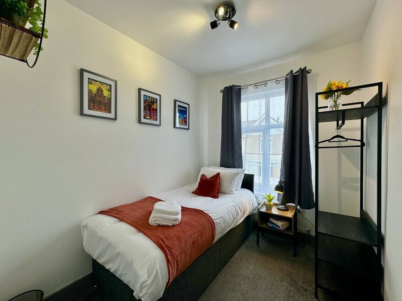 A single bed with a red blanket, a bedside table with a lamp, a small plant, and a wall-mounted shelf with a hanger.