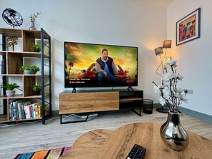 A living room with a TV, wooden entertainment unit, books, a vase with flowers, and a remote on a coffee table. - Lenhurst House – Contractors, Families, Holidays (Cleethorpes)