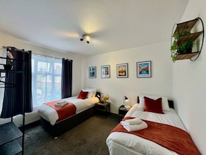 A hotel room with two beds, a window with curtains, a wall-mounted shelf with plants, and framed pictures on the wall. - Lenhurst House – Contractors, Families, Holidays (Cleethorpes)