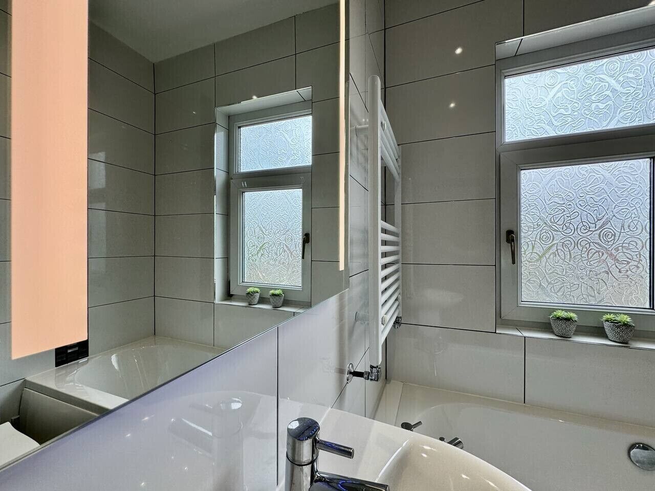 Et moderne badeværelse med et stort spejl, et badekar og to vinduer med mønstret glas.