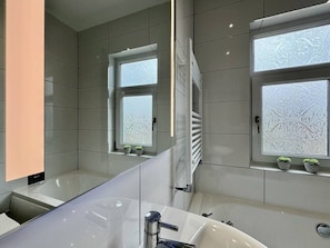 A modern bathroom with a large mirror, a bathtub, and two windows with patterned glass. - Lenhurst House – Contractors, Families, Holidays (Cleethorpes)
