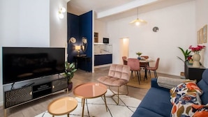 Ein modernes Wohnzimmer mit Flachbildfernseher, einer Couch, einem Esstisch mit Stühlen und einer Küchenzeile im Hintergrund.