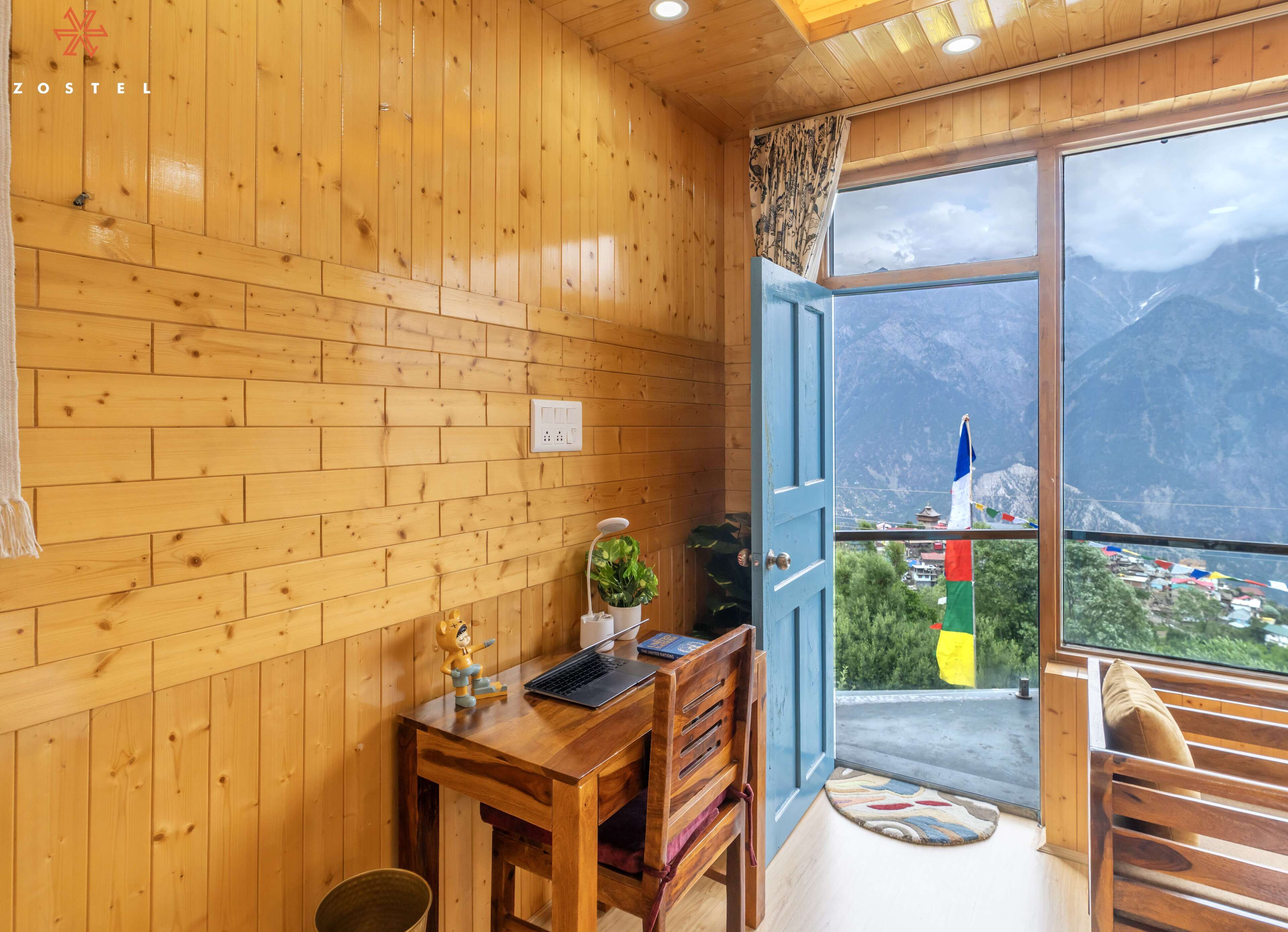A wooden room with a desk, a chair, a laptop, a plant, and a window with a mountain view.