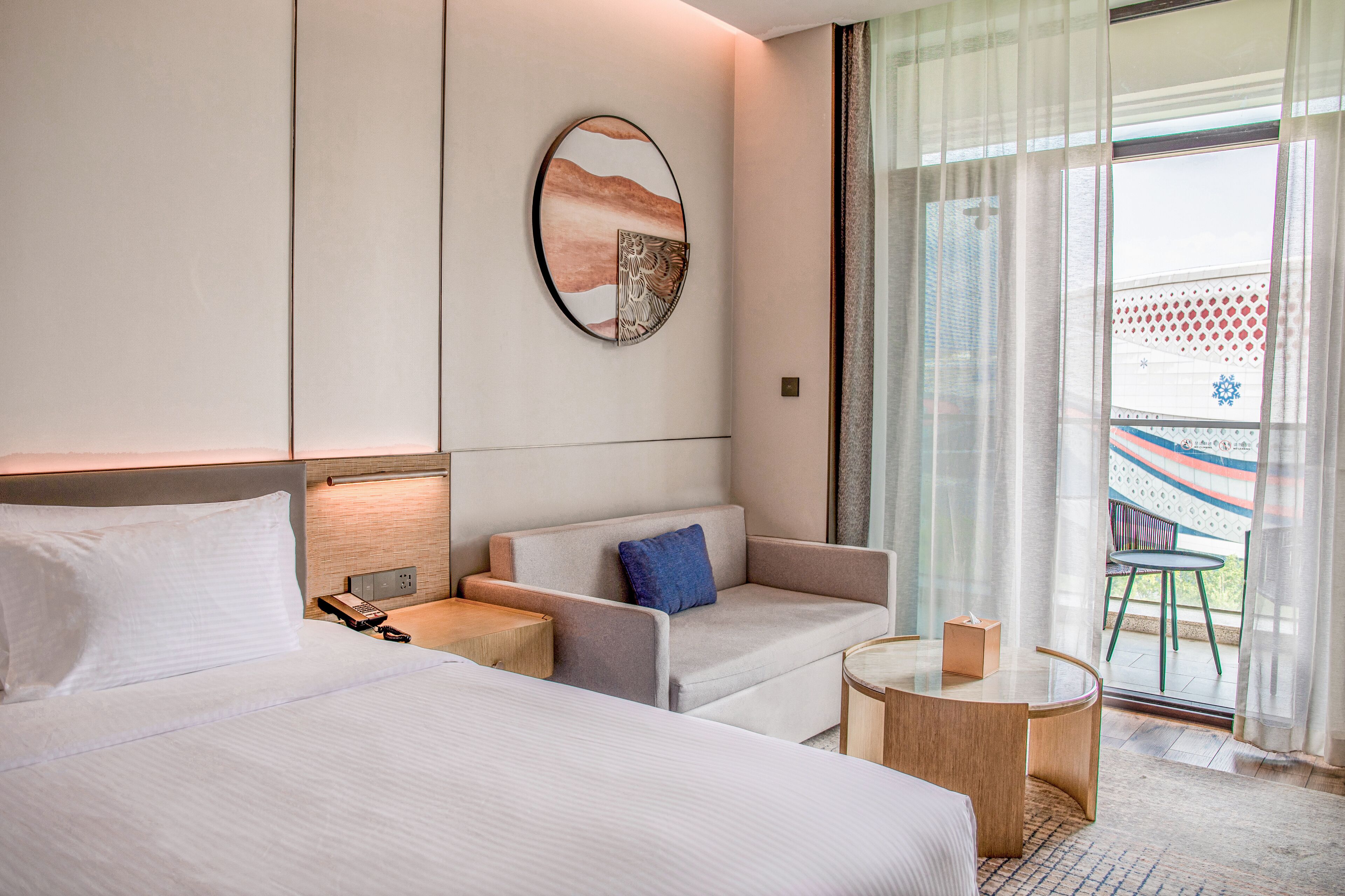 Quarto de hotel com uma cama, um sofá, uma mesinha e vista para o exterior através de uma grande janela.