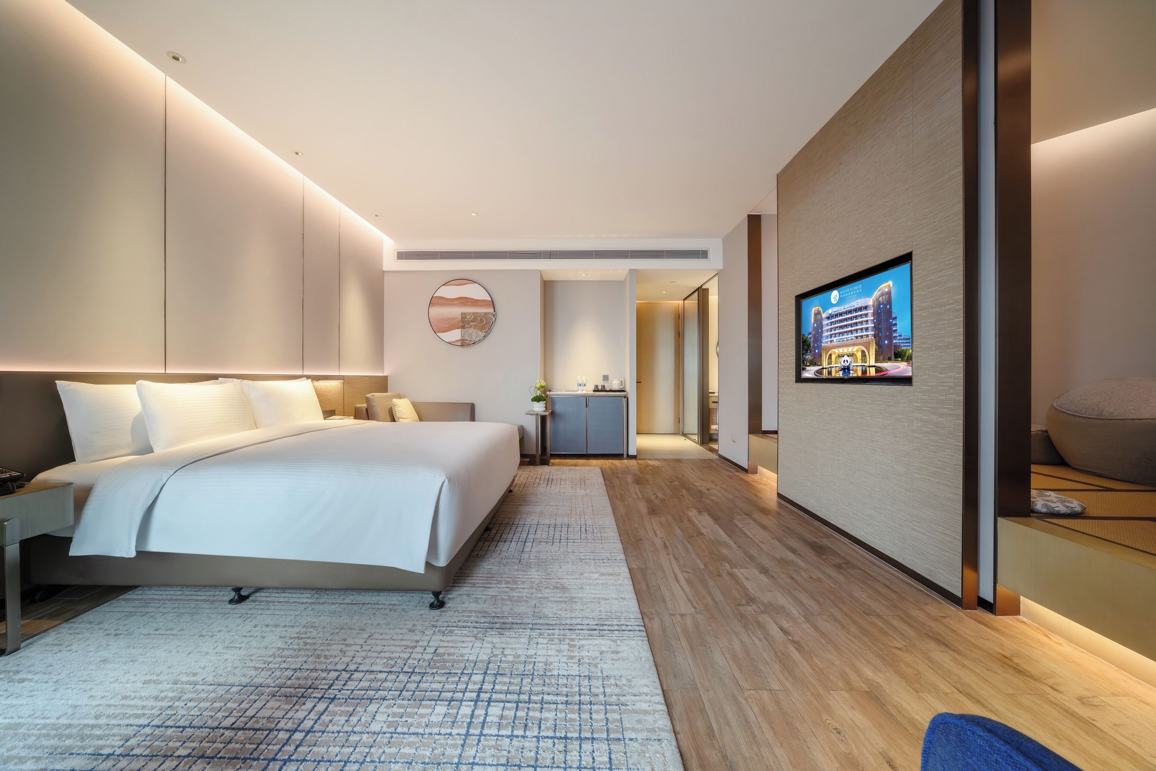 A modern hotel room with a large bed, a flat-screen TV mounted on the wall, a small kitchenette, and a bathroom visible through an open door.