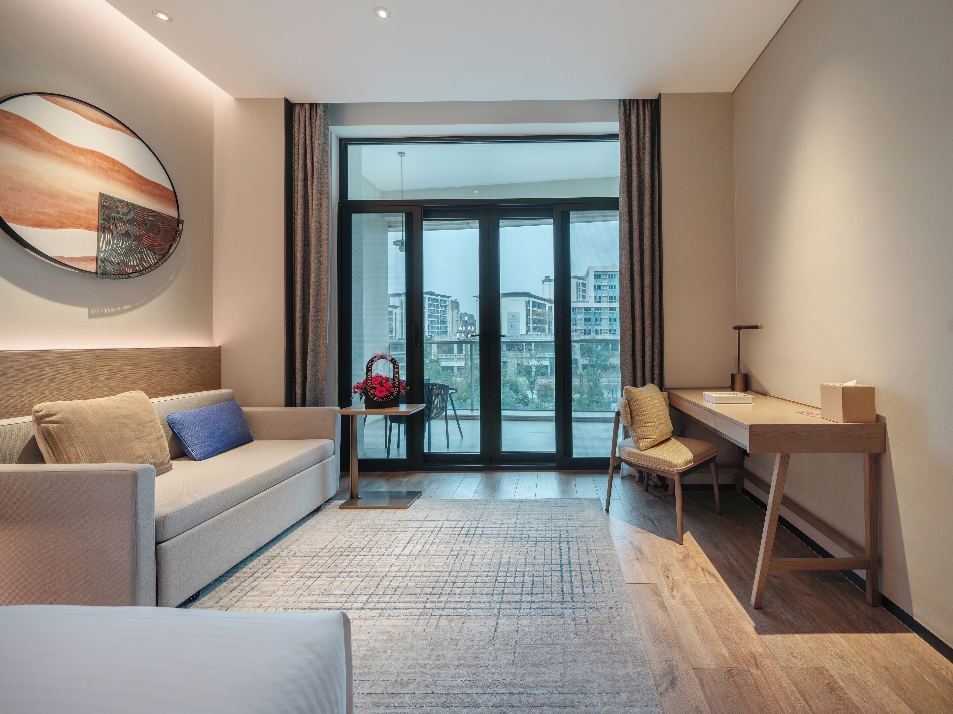 A modern hotel room with a sofa, a desk, a chair, and a large window offering a city view.