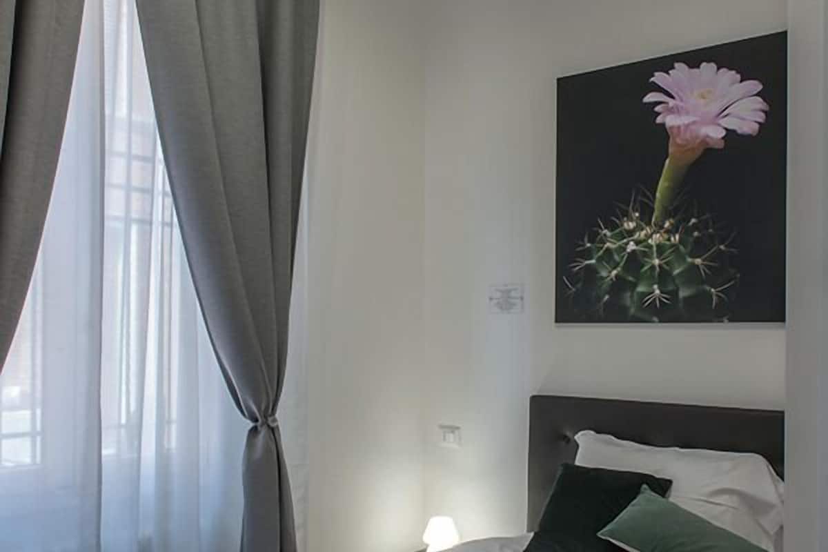 Ein Schlafzimmer mit Bett, einem Fenster mit Vorhängen, einem Nachttisch und einem an der Wand hängenden Bild.