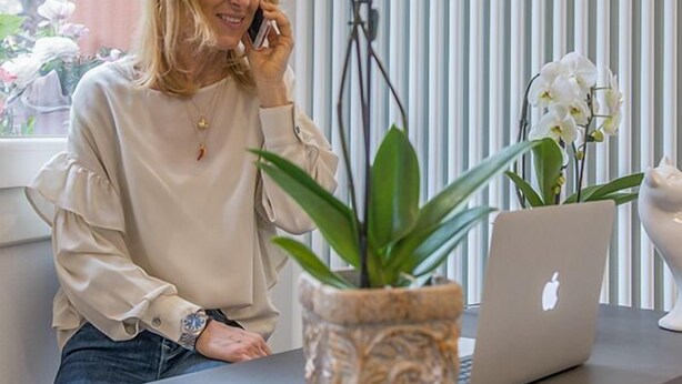 Une personne assise à un bureau, devant un ordinateur portable, avec une plante en pot et une fenêtre équipée de stores.