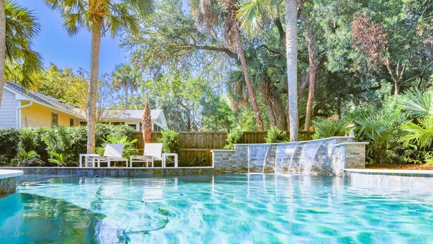 A swimming pool with a waterfall feature, surrounded by lush greenery and palm trees.