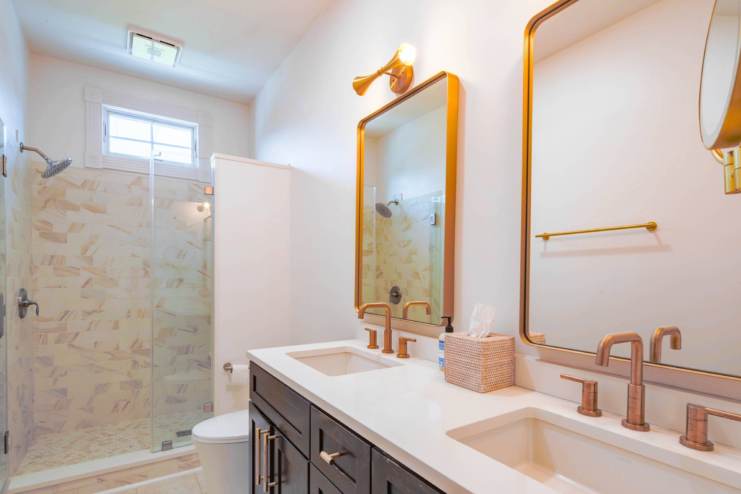 A bathroom with a glass shower enclosure, a white toilet, a double sink with gold fixtures, and a large mirror with a gold frame.