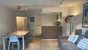 Un salon moderne avec un canapé gris, une table à manger avec des chaises, et une cuisine en arrière-plan.