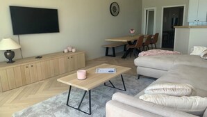 A modern living room with a sofa, a coffee table, a TV mounted on the wall, a dining area with chairs, and a kitchen area with cabinets. - Apartment near Port with Scenic Views (Nieuwpoort)