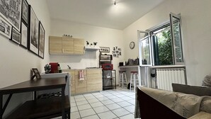 A compact kitchen with wooden cabinets, a gas stove, and a microwave. There are framed pictures on the wall, a window with a view of greenery, and a tiled floor.