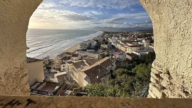 A coastal town viewed through a stone archway, featuring buildings, a beach, and a clear sky.