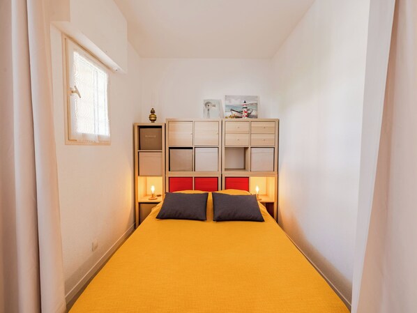 Une petite pièce comprenant un lit, une table de chevet avec une lampe, une fenêtre et un meuble de rangement intégré avec des tiroirs et des étagères.