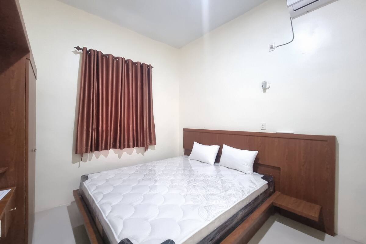 A bedroom with a wooden headboard, a bed with white bedding, a brown curtain, and a small wooden bench.