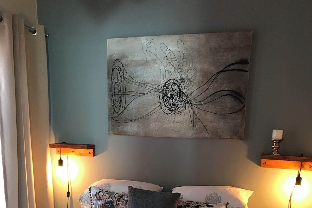 Una cama bien hecha con una colcha de estampado floral, dos almohadas y toallas enrolladas sobre la cama. Hay una obra de arte en la pared sobre la cama y dos lámparas de pared a cada lado.