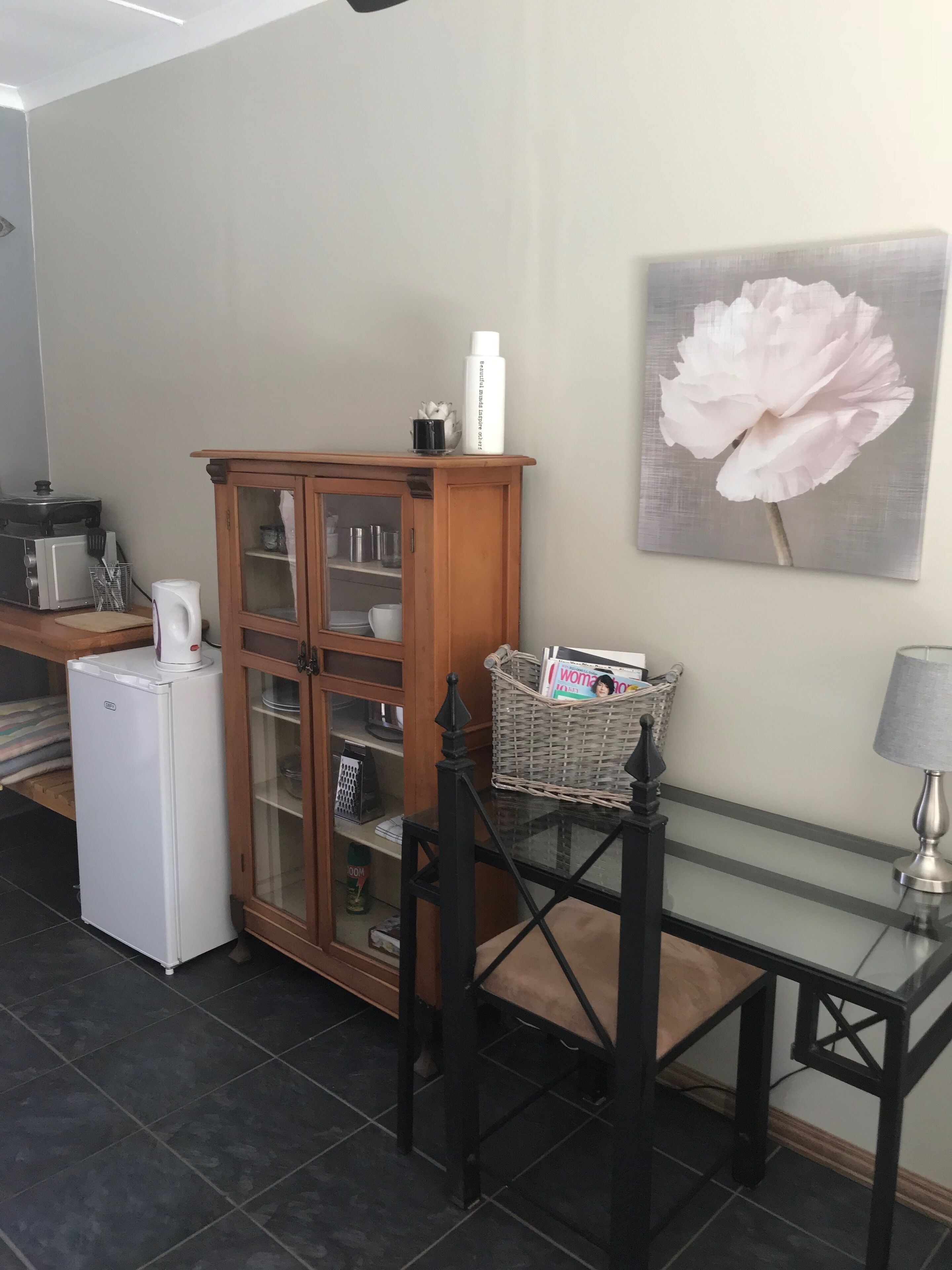 A kitchen with a refrigerator, microwave, and a cabinet with glass doors. There is a table with a lamp and a basket containing magazines.