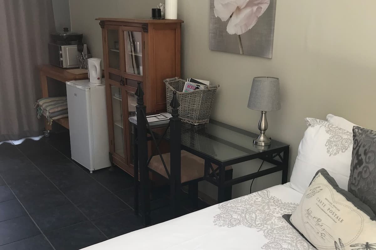 Un dormitorio con cama, mesita de noche, lámpara, silla, una pequeña mesa, refrigerador, microondas, armario y un cuadro de flores en la pared.