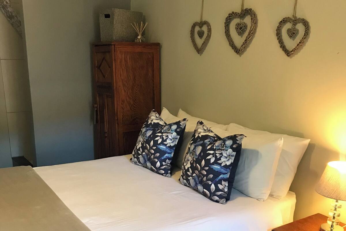 Una cama bien hecha con almohadas florales, una mesita de noche con lámpara y decoraciones de corazón en la pared.