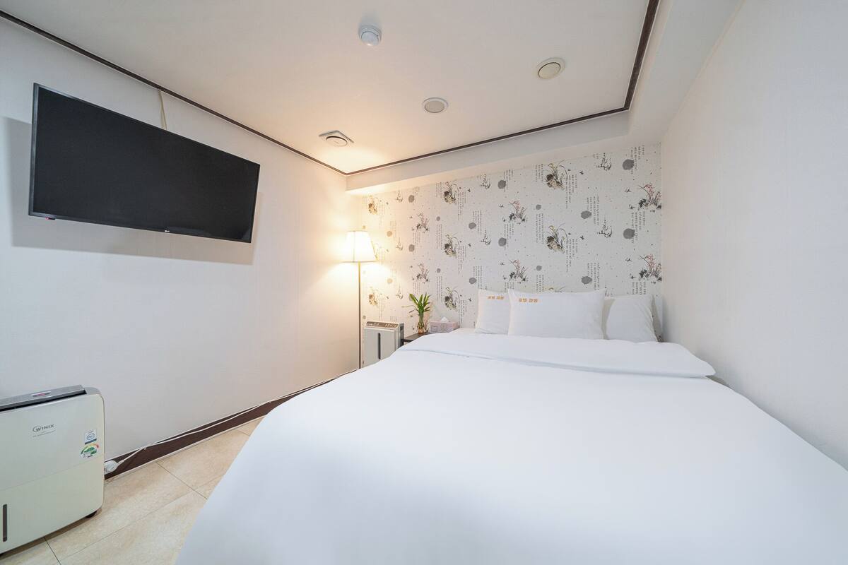 Hotellihuone, jossa on seinälle asennettu taulutelevisio, yöpöytä lampun kanssa ja valkoisilla lakanoiden kanssa varustettu sänky.