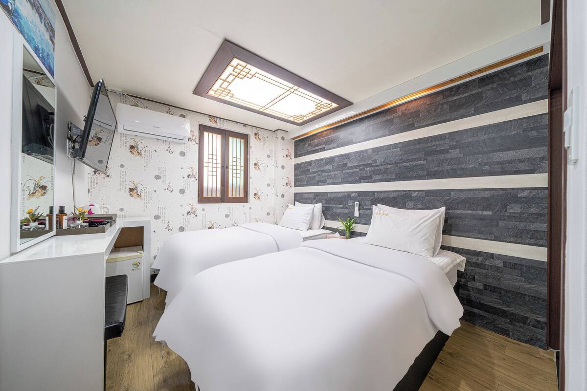 Hotellihuone, jossa on kaksi sänkyä, työpöytä, peili, ilmastointilaite ja kattovalaisin.