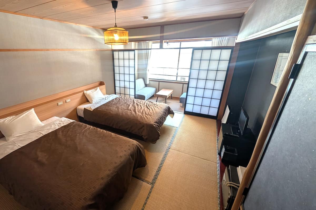 A hotel room with two beds, a tatami mat floor, a traditional Japanese sliding door, and a small table with a chair.