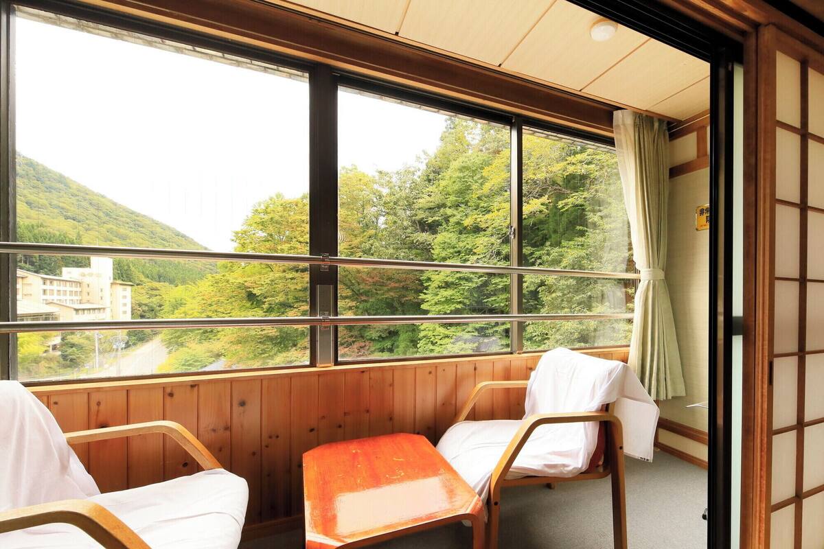 A room with two wooden chairs and a small table, large windows with a view of a forested hill and buildings, and a traditional sliding door.