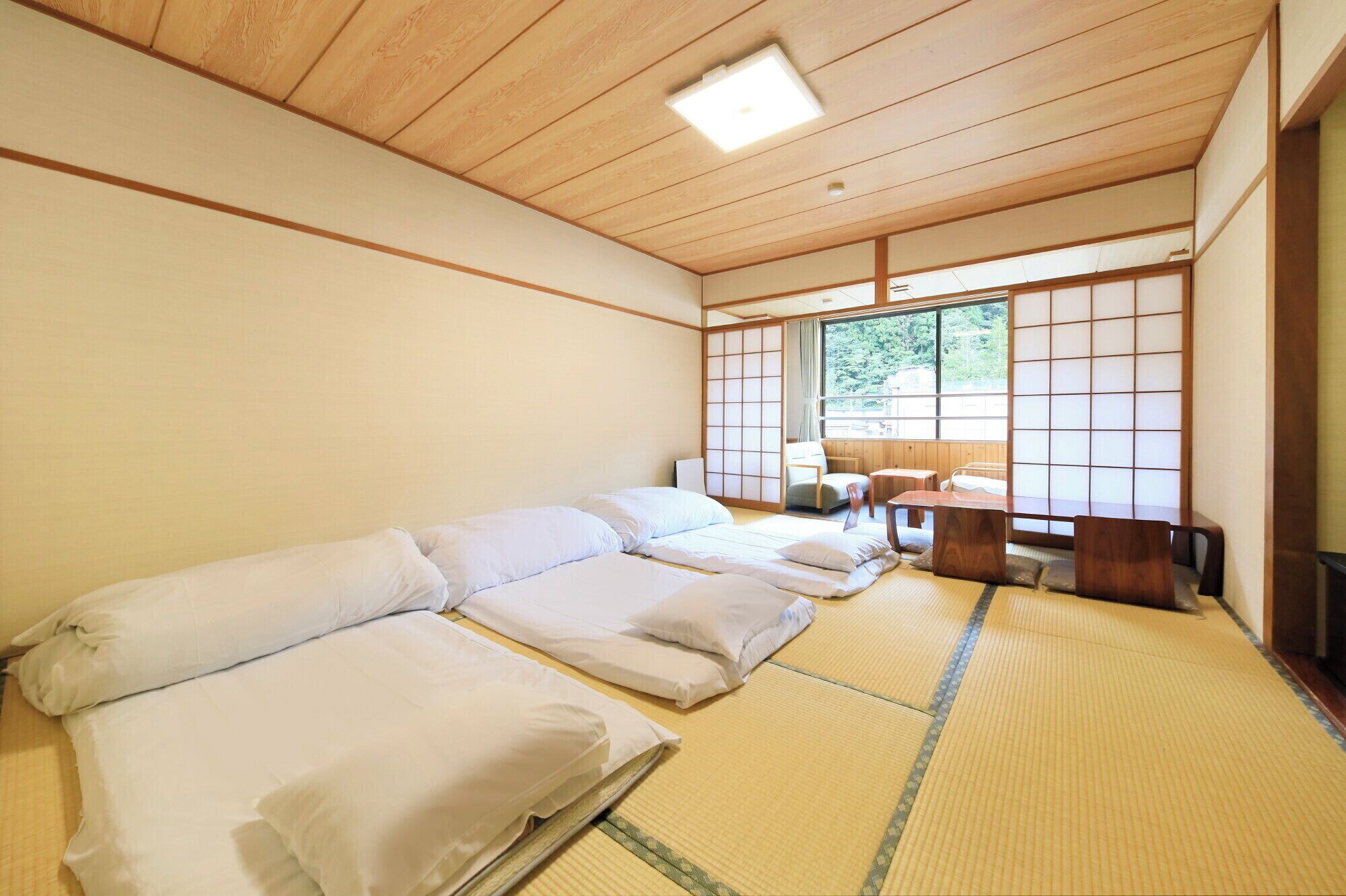 Una habitación tradicional japonesa con esteras de tatami, una cama de madera baja, una mesita y vistas al verdor exterior.