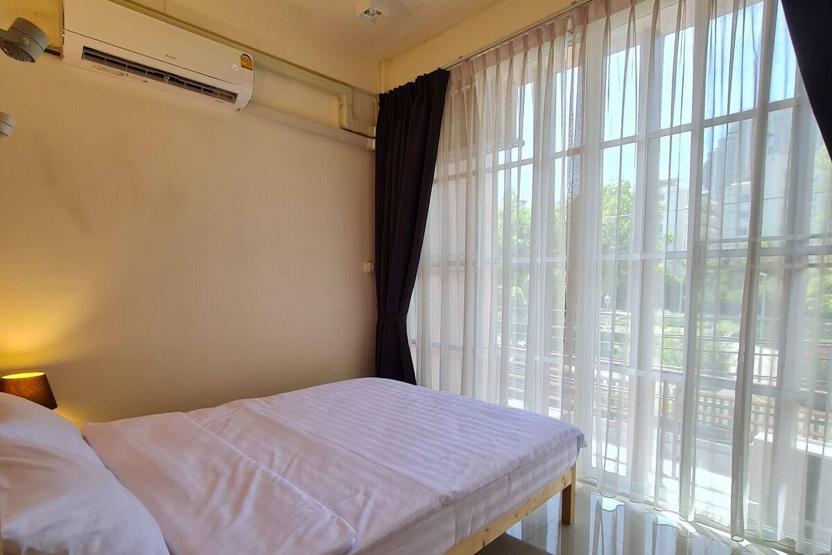 A room with a bed, a wooden nightstand, a window with sheer curtains, and an air conditioning unit on the wall.