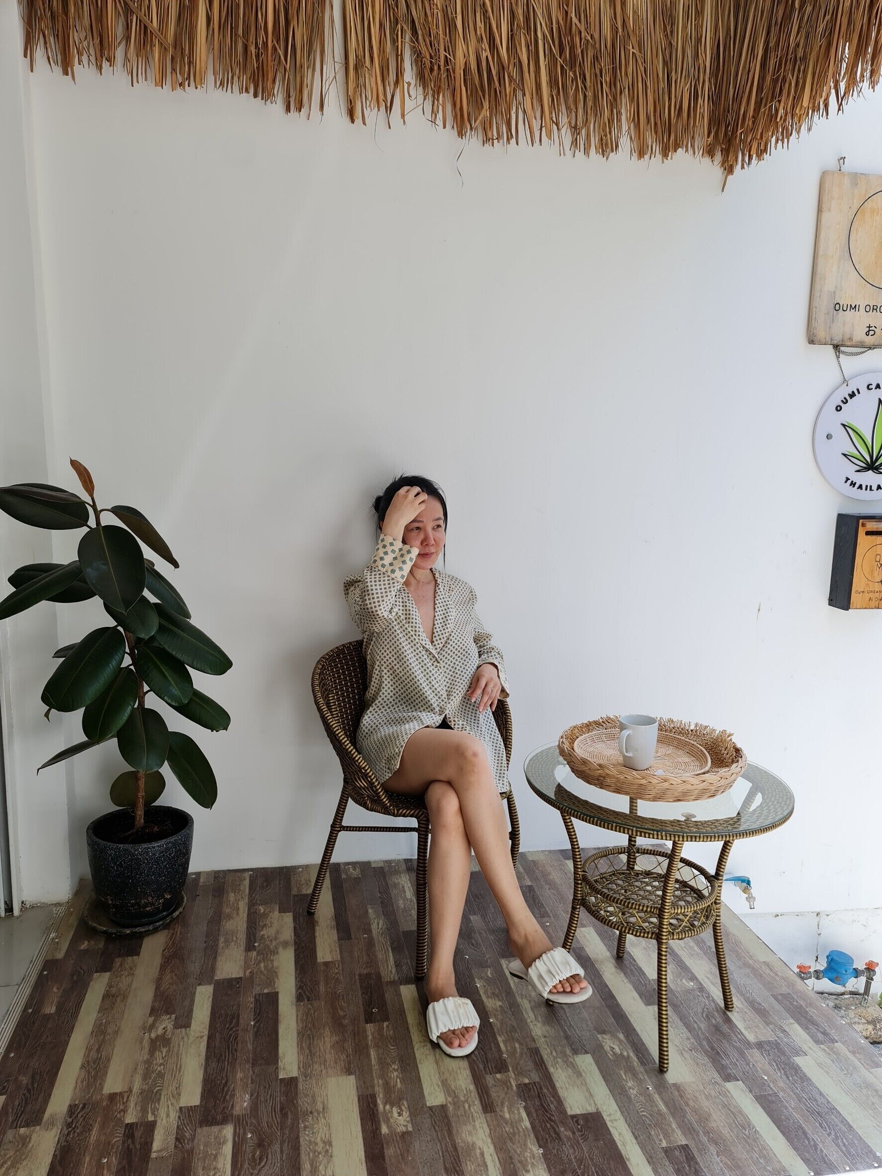 A person sitting on a wicker chair in a room with a wooden floor, a potted plant, and a small round table with a cup.
