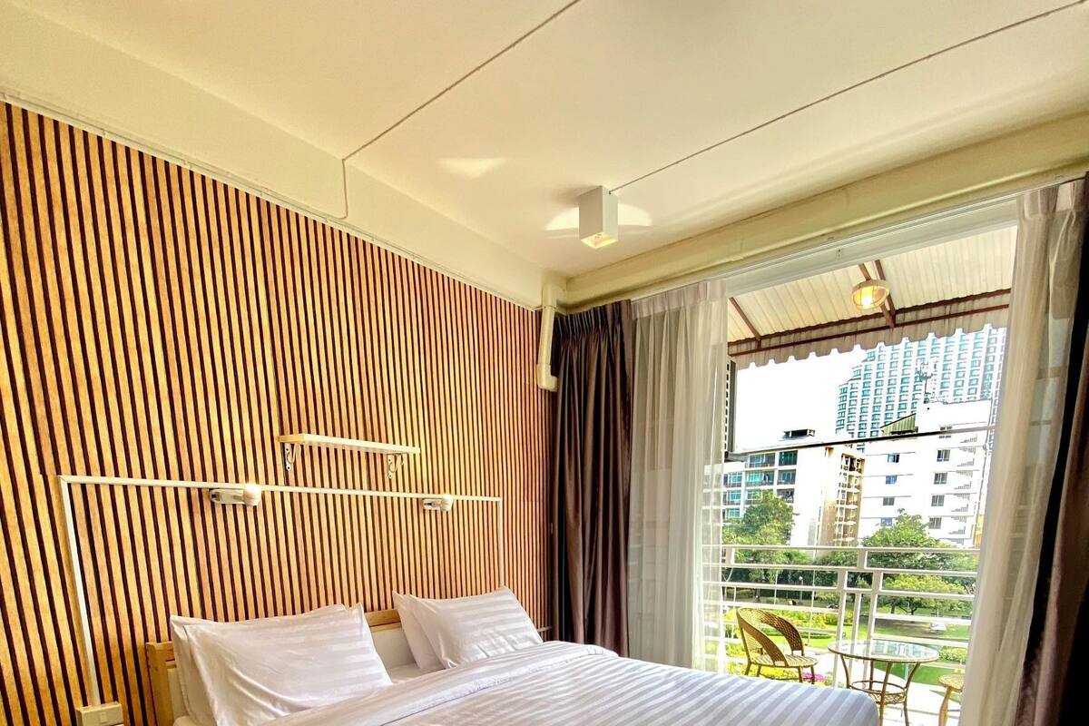 A hotel room with a large bed, a bedside table, a wooden slatted wall, and a view of buildings through a window.
