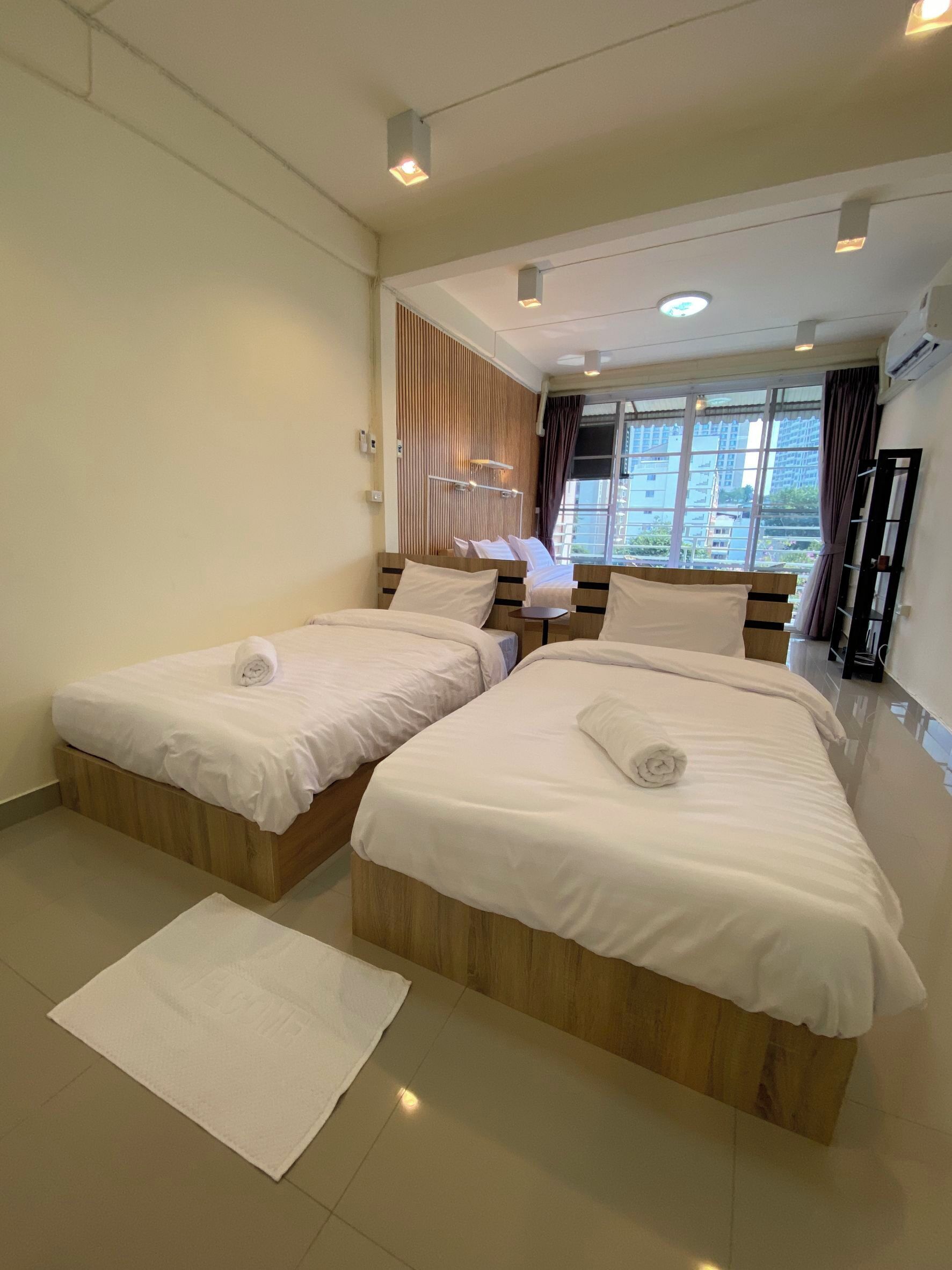 A hotel room with two beds, a small table, a window with a view of buildings, and a ceiling with recessed lighting.
