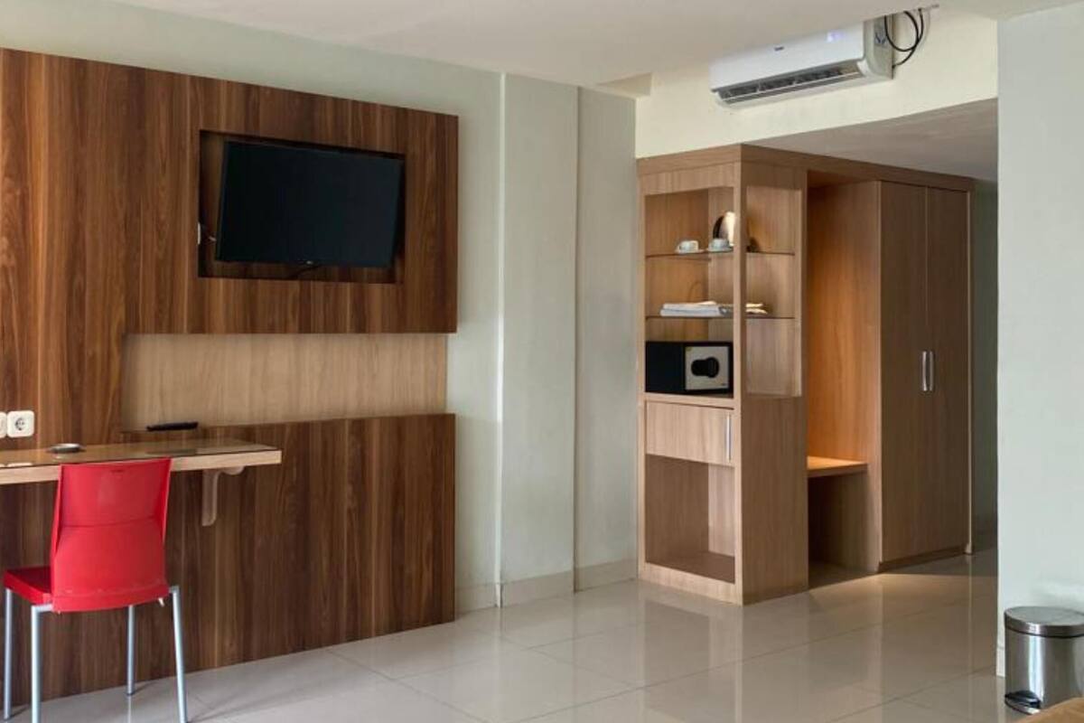 Ein modernes Hotelzimmer mit einem Holztisch, einem roten Sessel, einem Bett mit brauner Bettdecke und einem Schrank mit integrierten Regalen.